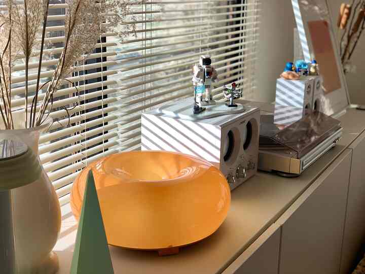 暖かい日差しが差し込む白を基調としたリビング、ブラインドとオーディオ機器、オレンジ色の装飾品が配置された雰囲気のあるインテリア