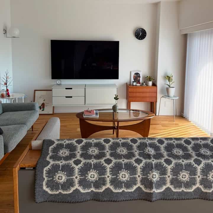 ナチュラルと白を基調とした北欧風リビングルーム、中央にオーバルの木製ガラスコーヒーテーブル、グレーのソファ、ミナペルホネンのブランケットが配された印象的な空間