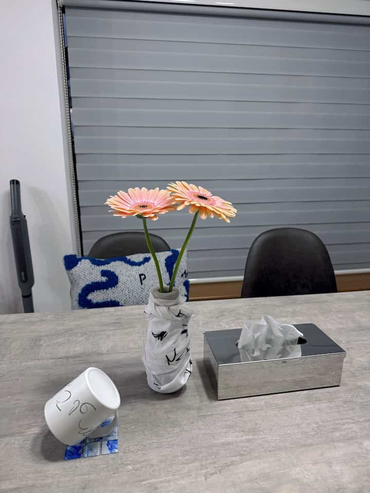 グレーのブラインドを背景にしたダイニングルーム、ナチュラルカラーのダイニングテーブルに花瓶と花、マグカップが配置された雰囲気のある空間