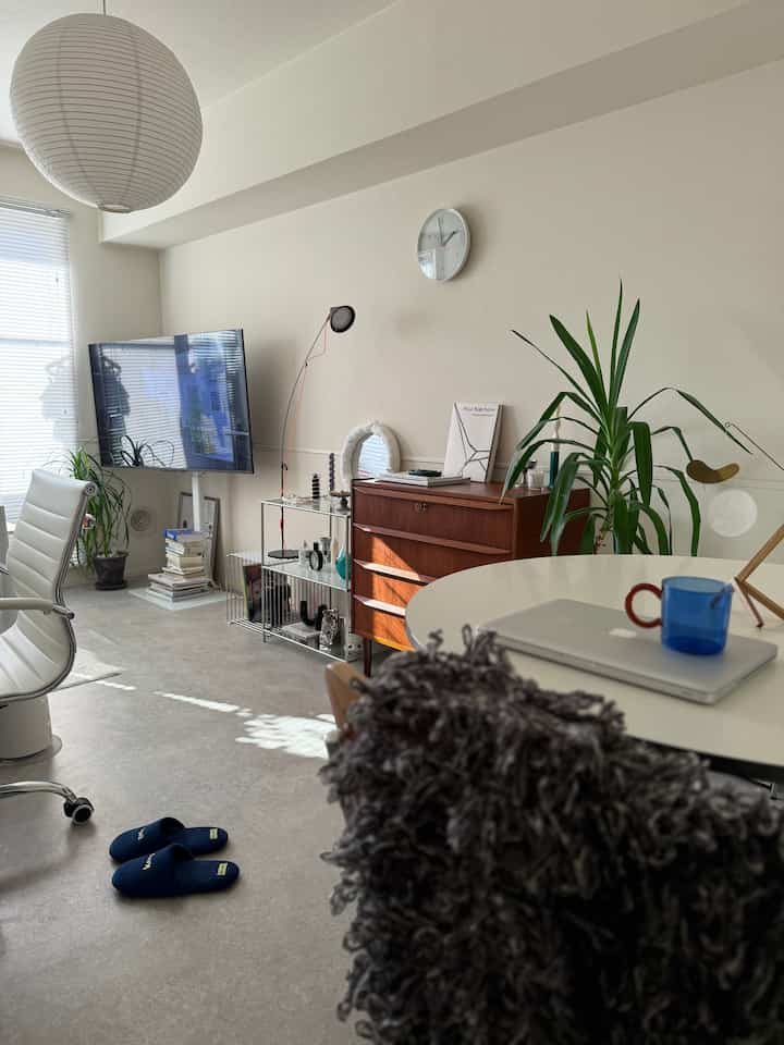 Mid-century modern long and narrow living and dining room with white walls, natural wood dresser, sofa, dining table, and large green plant