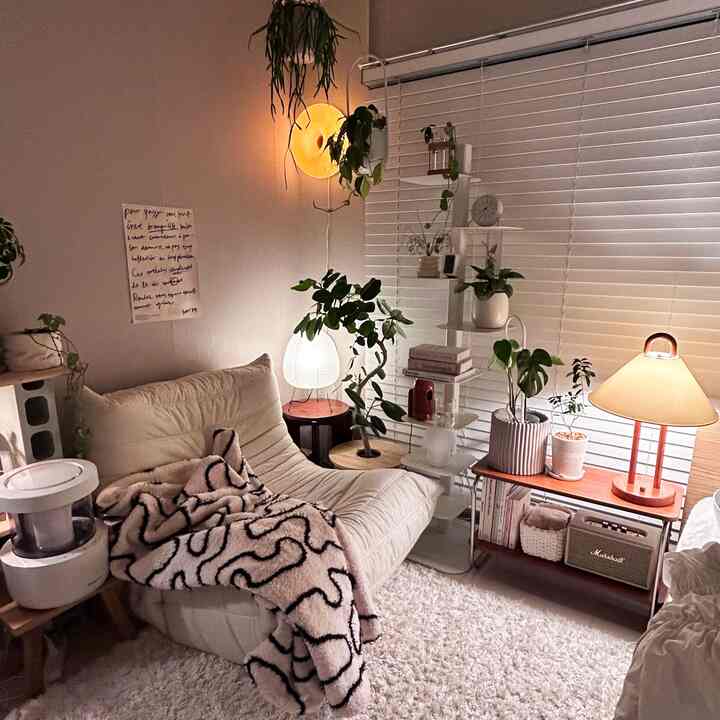白とベージュ調のリビング、快適なソファとブランケット、複数の植物と暖かい照明が配置された自然でシンプルな雰囲気のインテリア