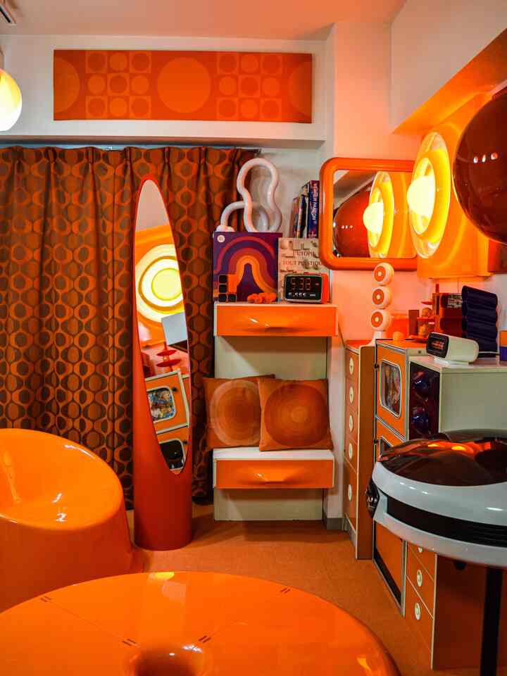オレンジ色調のレトロな小部屋で、丸みのある家具とペンダントライト、鏡が配置された温かみのあるインテリア