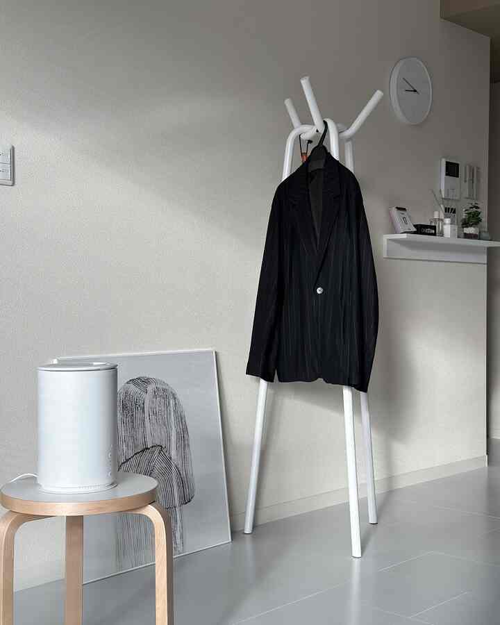 Modern living room in white and monotone tones, featuring a humidifier on a stool, coat rack with clothing, and a wall clock arranged neatly