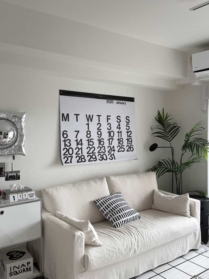 White and gray toned living room featuring a cream sofa, modern wall calendar, and greenery in a simple, clean setting