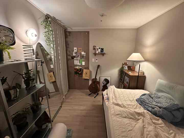 柔らかなベージュ色調の小さなワンルームの寝室、窓辺に植物スタンドと壁付け照明、右側にベッドとフロアランプが暖かい雰囲気を演出しています。