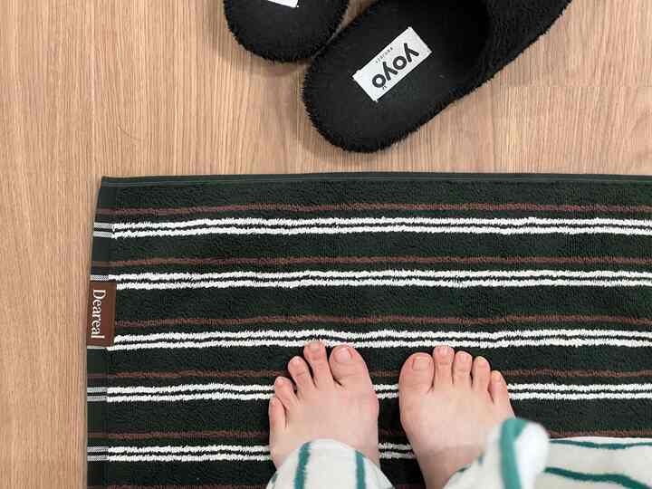 ブラウンの木目調フロアに縞模様のバスマットと黒い室内スリッパが置かれたバスルームのインテリア