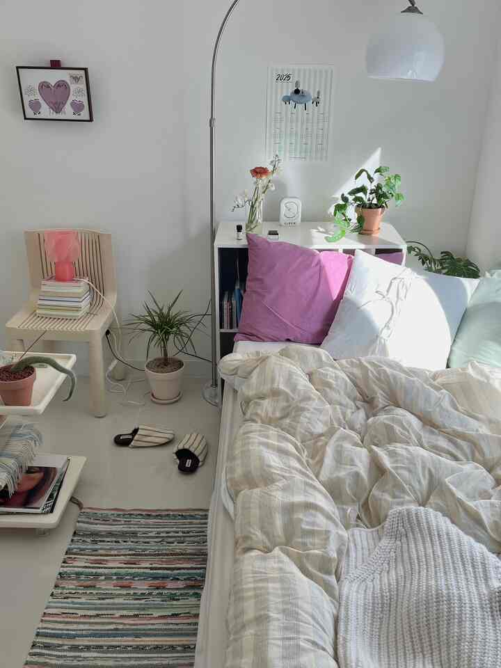 白を基調とした小さなワンルーム寝室、枕や寝具、植物、小さな収納棚が配置された雰囲気のあるインテリア