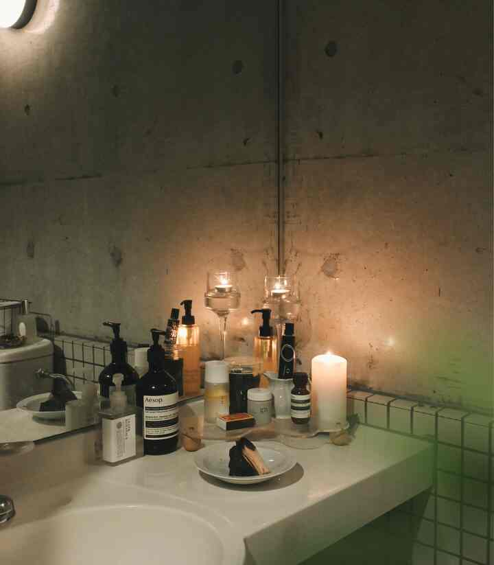 Natural-toned bathroom with gray concrete wall and tiled sink, featuring various candles and diffusers creating a cozy atmosphere