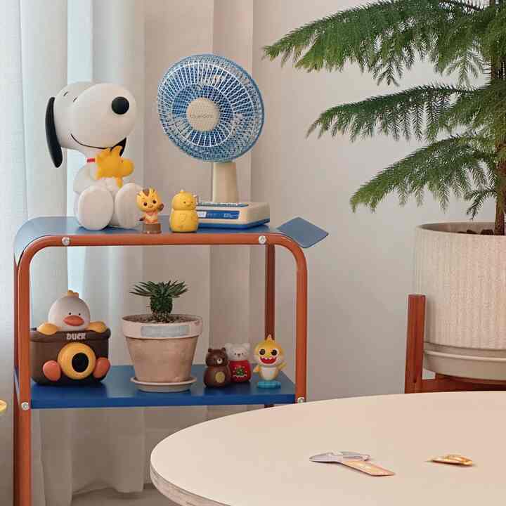 白い壁とカーテンのリビングの一角に、ブルーのサイドテーブル上にカラフルな子供の玩具や小さな植物、青い扇風機と大きなナチュラルな観葉植物が配置された雰囲気のあるインテリア