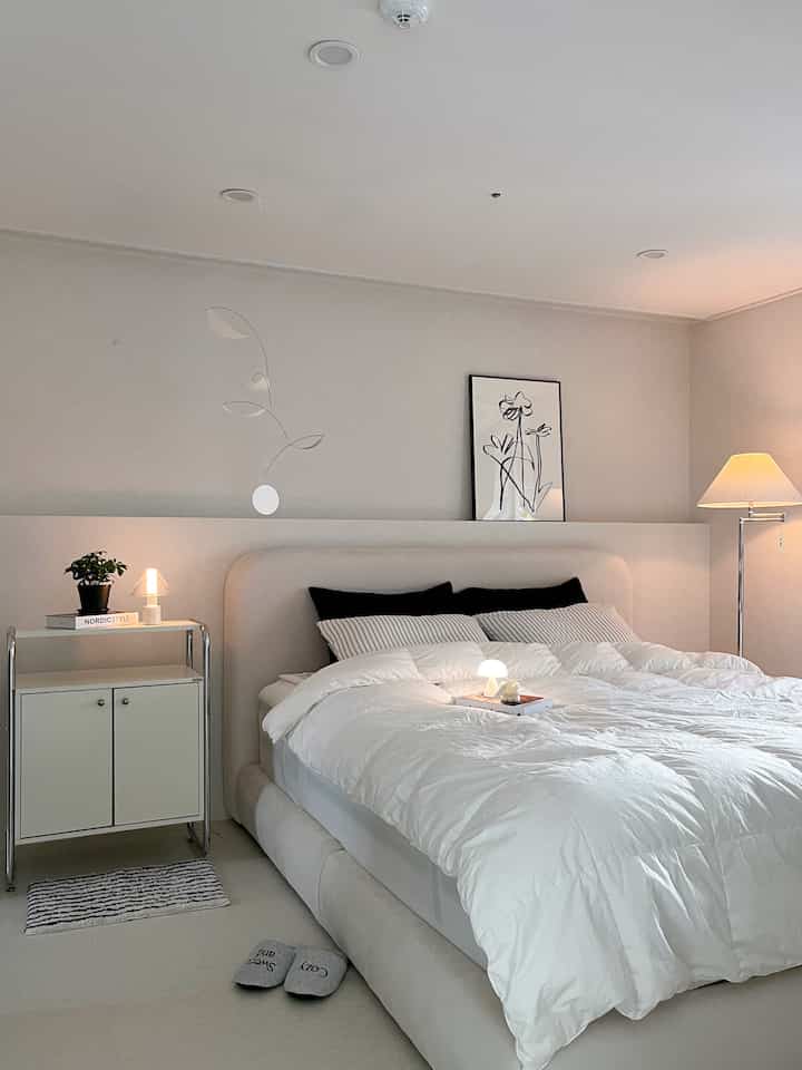 White and beige toned bedroom featuring a cozy bed, poster on wall, and floor lamp creating a simple and warm atmosphere