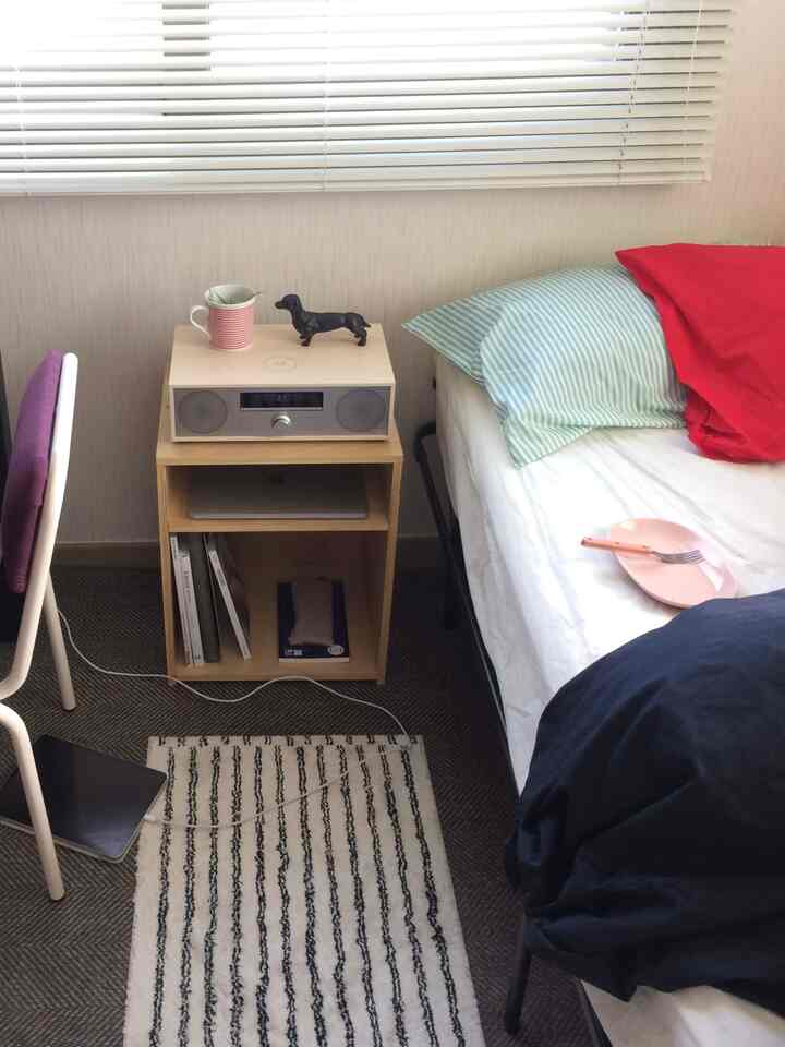 白い壁とブラインド、青い寝具、木製のナイトテーブルが配置されたコンパクトな一人暮らしの寝室