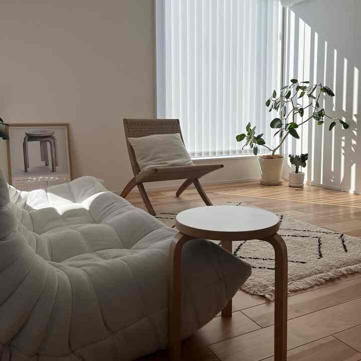 Natural-toned living room featuring a white sofa, wooden stool, armchair, and large plant by the window with bright sunlight