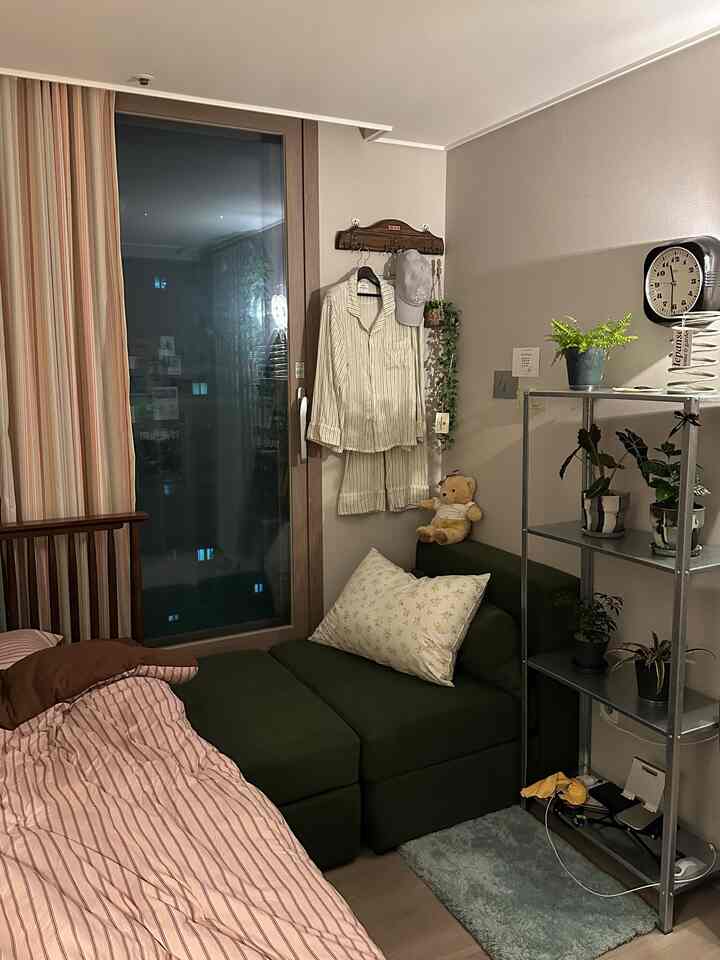 暖かいベージュと木目調の調和が感じられる小さな寝室、ウォールナットベッドと緑の1人用ソファ、植物と壁掛け時計が配置された雰囲気のあるインテリア