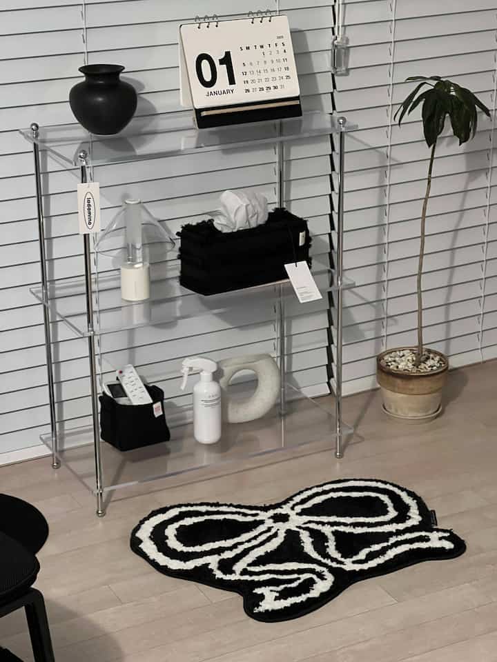 Monotone living room featuring a black-and-white bow-shaped mini rug, transparent acrylic shelf, wood-tone flooring, and an indoor plant for a clean aesthetic