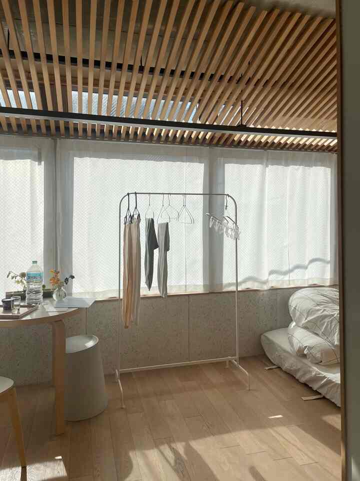 ナチュラルな木目調と白いカーテンが調和するコンパクトな6畳の寝室、洋服掛けラックと小さなスツールが配置された落ち着いた空間