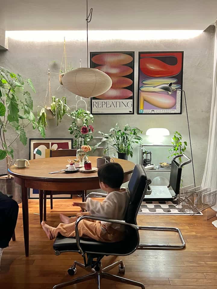 Wood-tone and white living room featuring round dining table, office chair, and various plants with a warm atmosphere