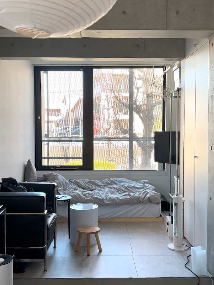 Bright white and natural toned compact single household bedroom featuring a large window and modern black sofa with clean aesthetic