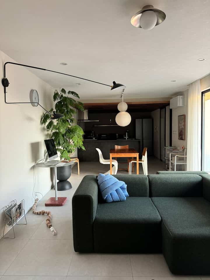 White walls and gray floors define a mid-century modern living room featuring a dark green sofa and wooden dining table in a natural atmosphere