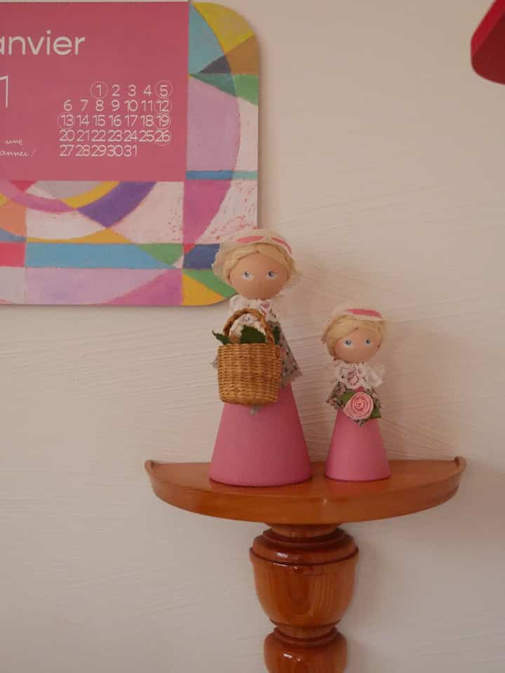 暖かみのある木目調のコーナー棚に置かれたピンクのドレスを着たヴィンテージの木製人形と、壁のカラフルなカレンダーアートのある空間