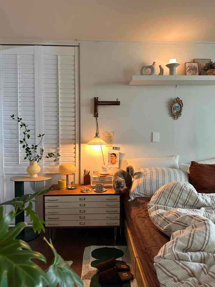 Cozy bedroom in warm brown and white tones featuring a bed, side table, wall-mounted lamp, and a cat on the bedside table