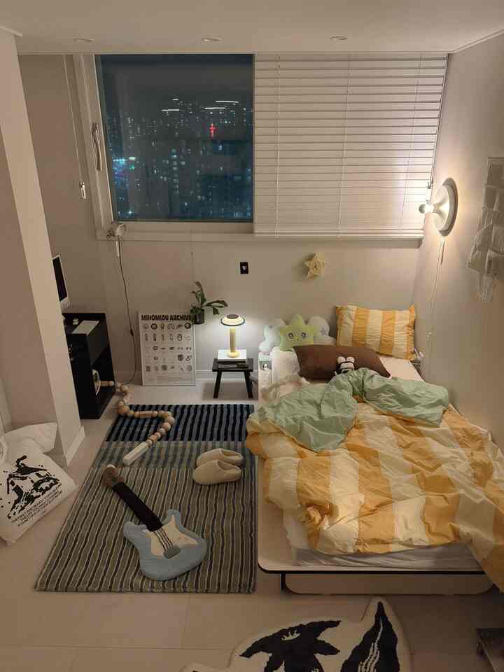 黄色の寝具が置かれた小さなワンルームの寝室、白い壁と木目調の家具が温かみのある雰囲気を作るインテリア