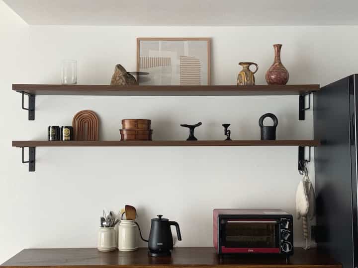 白い壁とナチュラルなブラウンの木製棚が配置された清潔感のあるキッチン、シンプルな装飾と調理器具が調和した雰囲気