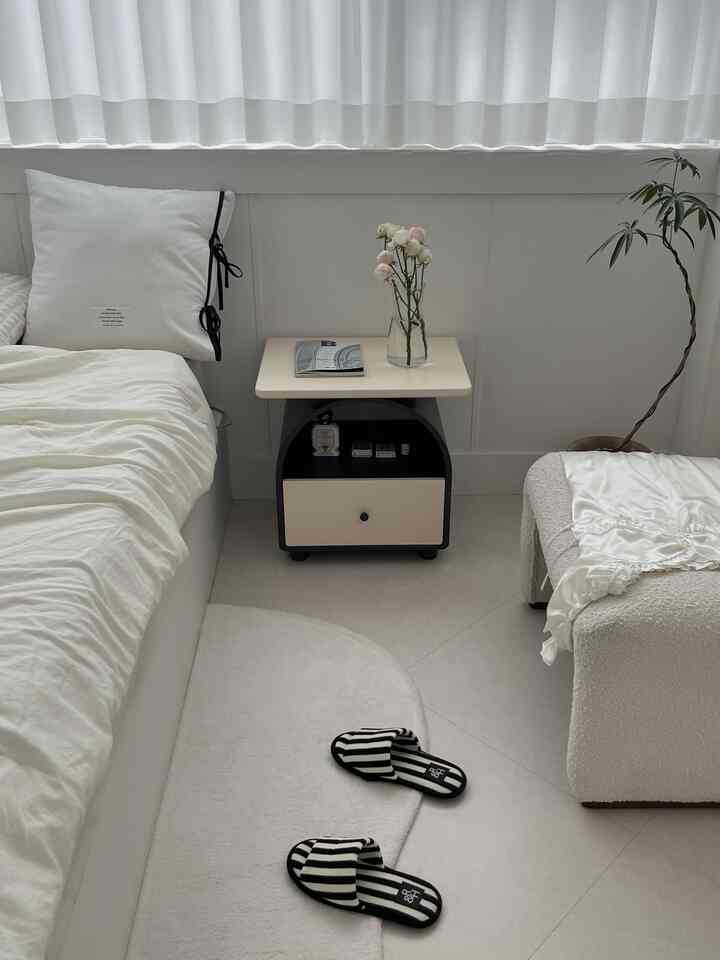 ホワイトとブラックの色調の寝室、ベッドの横に黒いナイトテーブルと花瓶、柔らかいラグとスリッパが配置された雰囲気のあるインテリア