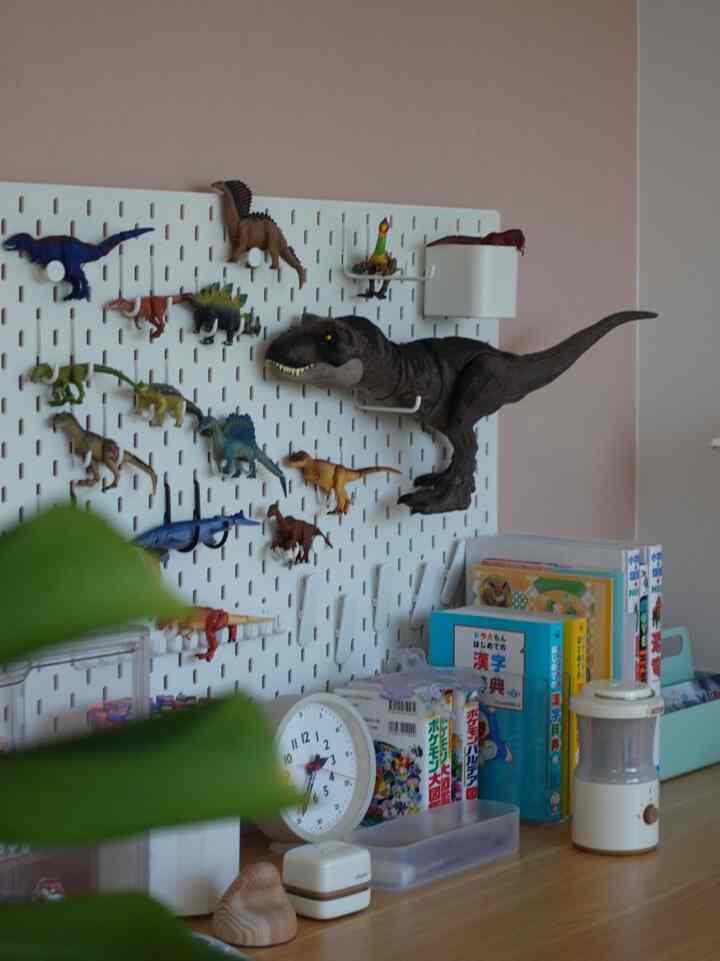 ピンク色の子供部屋に白いペグボードが設置され、恐竜のおもちゃや時計、本が木製の机の上に並べられた雰囲気のあるインテリア