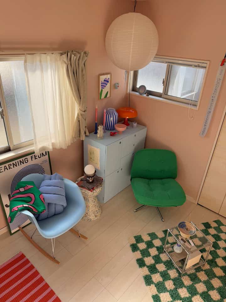 Pink-toned walls and natural wood floor define a cozy 6-tatami small room featuring a green armchair and blue rocking chair in a home office setup