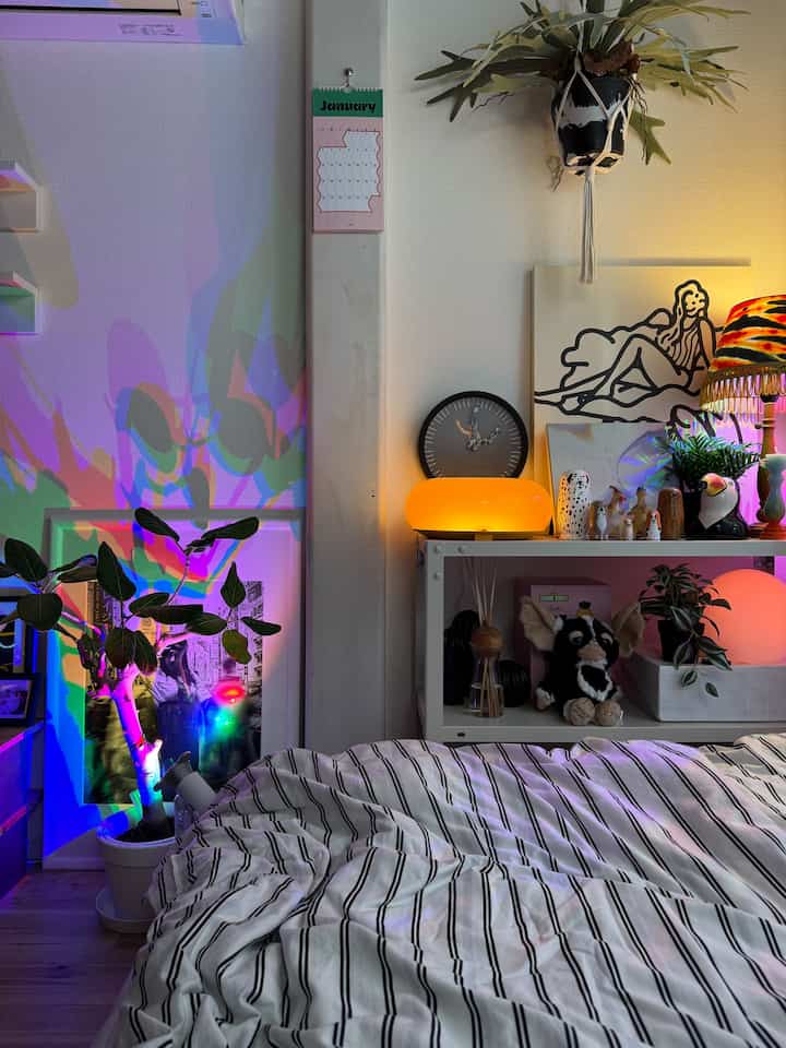 白と黒のストライプの寝具があるワンルームの寝室、多彩な間接照明が壁と植物を照らす雰囲気のあるインテリア