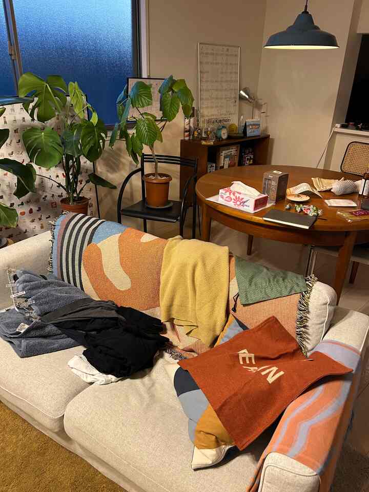 ナチュラルなブラウン調のリビング、ベージュの3人掛けソファと木製のダイニングテーブル、多彩なスローと植物が配された散らかった空間