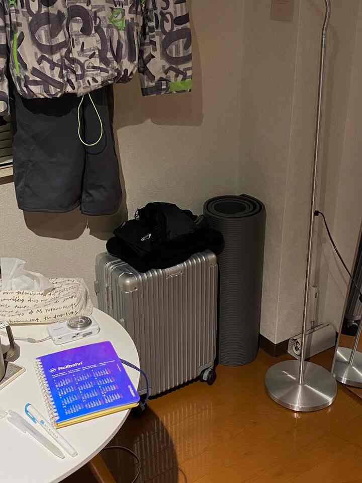 ブラウンの木製床とナチュラルな色調の壁があるコンパクトな一人暮らしの部屋。中央右にはシルバーのスーツケースとヨガマット、左には白い丸テーブルとブラインドが配置された印象的な空間