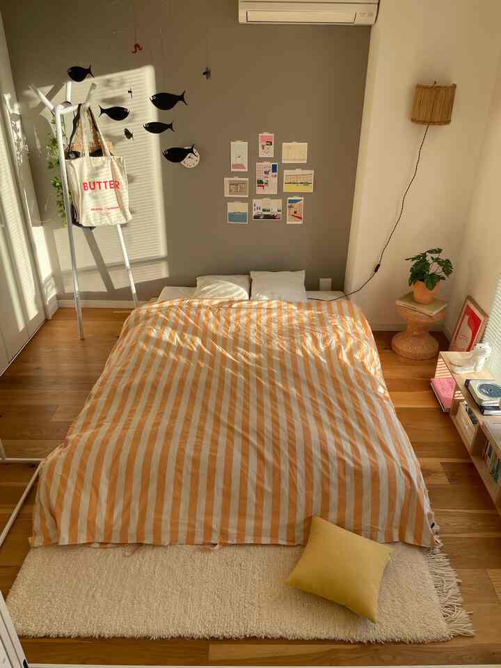 ナチュラルな木目調の寝室、オレンジのストライプ布団と白い壁照明が配置されたシンプルなインテリア