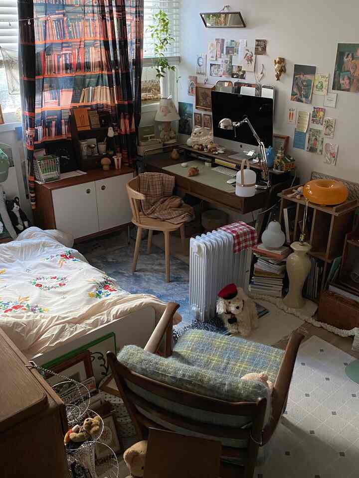 ナチュラルウッドトーンと白い壁の寝室兼ホームオフィス、書棚柄のカーテンとかわいい小物が配された居心地の良いインテリア