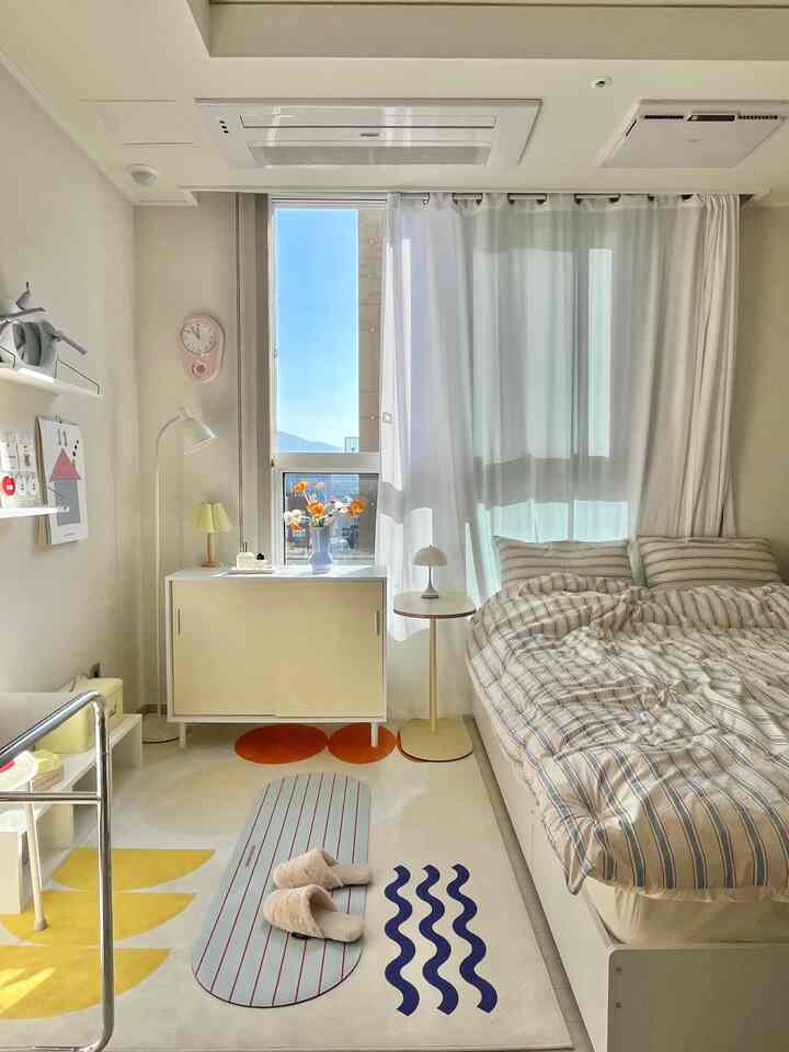 Bright white-toned studio bedroom with yellow accents, featuring a large window and geometric rug in a natural modern space