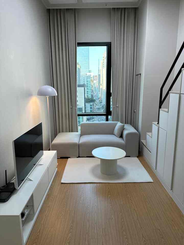 白とナチュラルウッドの調和が美しいメゾネットスタジオリビング、モダンなソファとコーヒーテーブル、収納付き階段が配置された印象的なインテリア