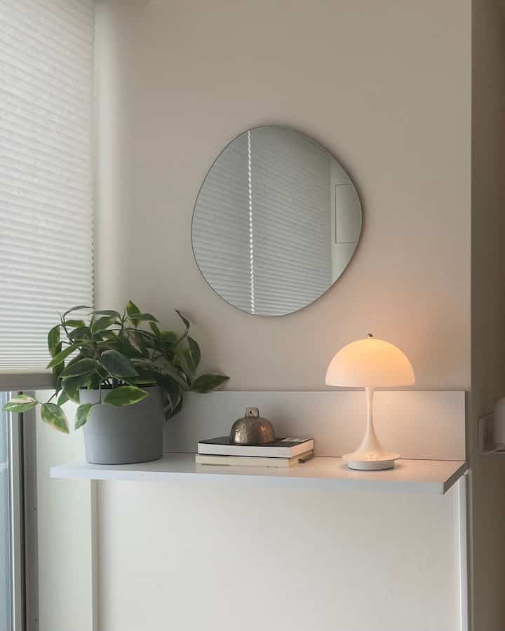 Minimal studio apartment corner with white wall-mounted shelf, green plant, round mirror, and warm table lamp creating cozy vibe
