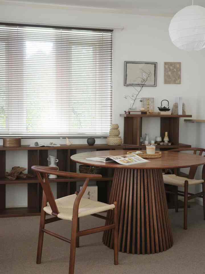 Warm wood tones in a natural modern dining room featuring a round wooden dining table, blinds-covered window, and wall shelving creating a cozy atmosphere