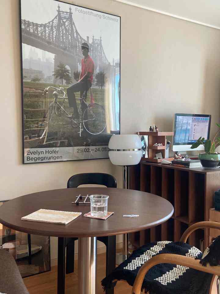 Living room with white walls and a brown round table, featuring a large poster, floor lamp, vanity shelving, and a desk creating a cozy interior