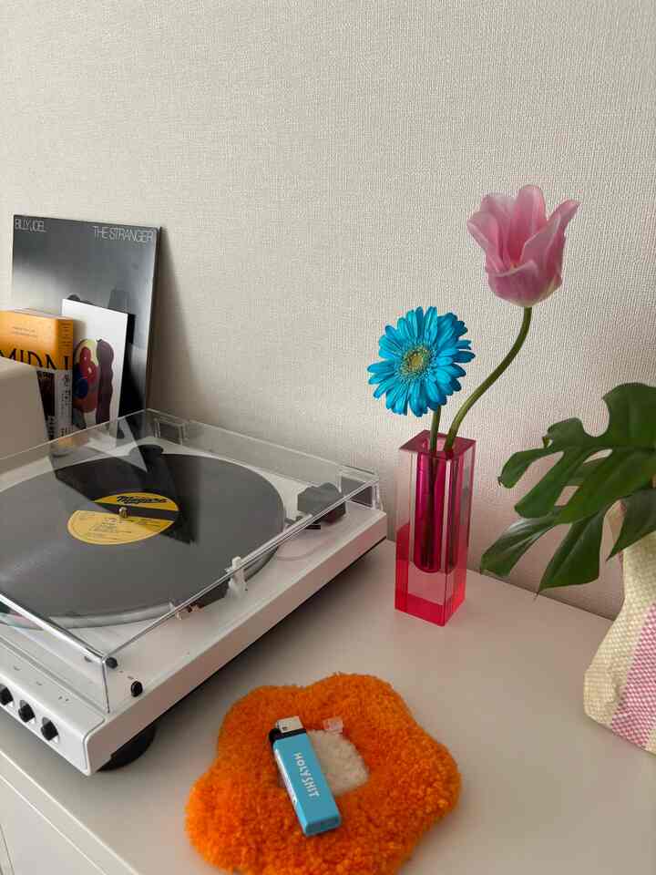 白を基調とした小さな空間に、白いレコードプレーヤーと透明なピンクの花瓶、青とピンクの花が置かれたミニマルなインテリア