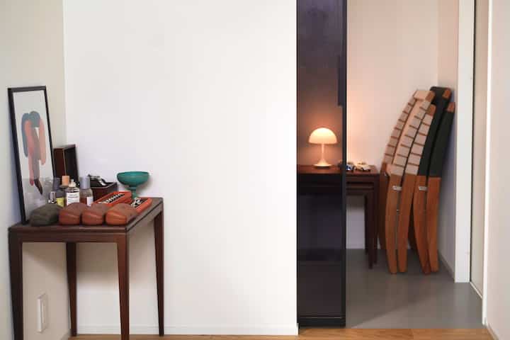 ナチュラルな色調とブラウンの家具が配された狭い玄関、サイドテーブルと折りたたみ椅子が調和する印象的な空間