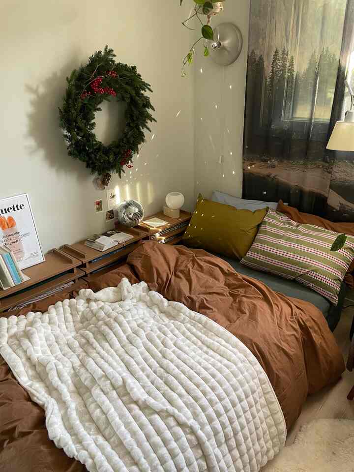 暖かみのあるブラウンとナチュラルな色調の寝室、柔らかな寝具と壁に掛けられたクリスマスリースが配置された雰囲気のあるインテリア