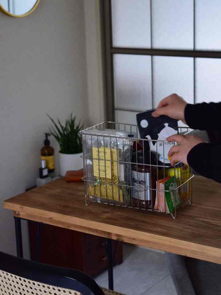 Natural wood tone dining table featuring a stainless steel wire basket organizing various tea and coffee packets in a tidy kitchen storage space