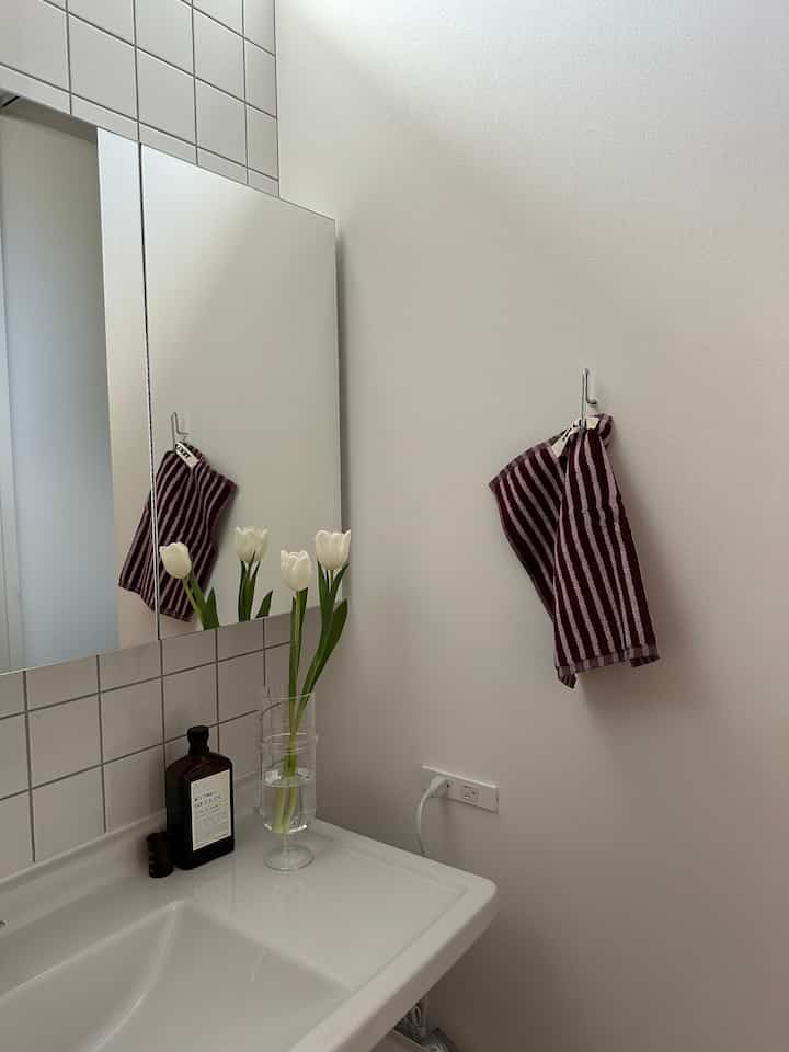 白を基調としたバスルーム、白い洗面台と鏡の上にチューリップが入った花瓶が置かれた清潔感のあるインテリア