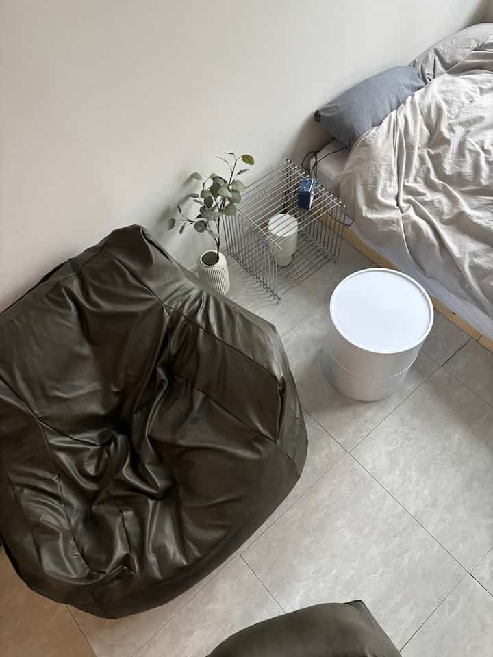Calm monotone bedroom-living space featuring a dark olive bean bag sofa and white stool with a simple, minimalistic decor