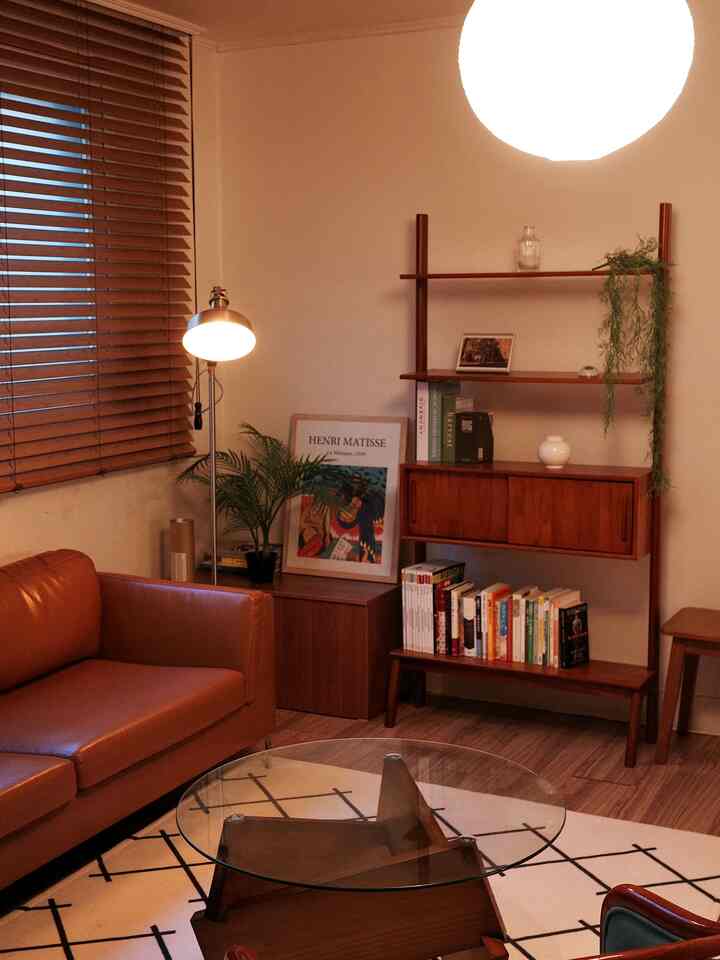 暖かいウッドトーンとブラウン家具が配置された居心地の良いリビングルーム、レザーソファと丸いガラスのテーブル、棚、電球照明が特徴的です。