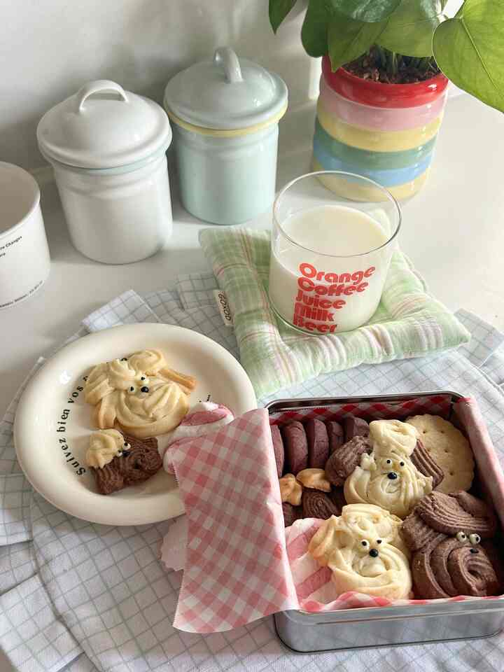 パステル調のダイニングテーブルに犬の形のクッキーとクッキー缶、牛乳のグラス、多色の植木鉢が配置された雰囲気のあるおうちカフェ空間