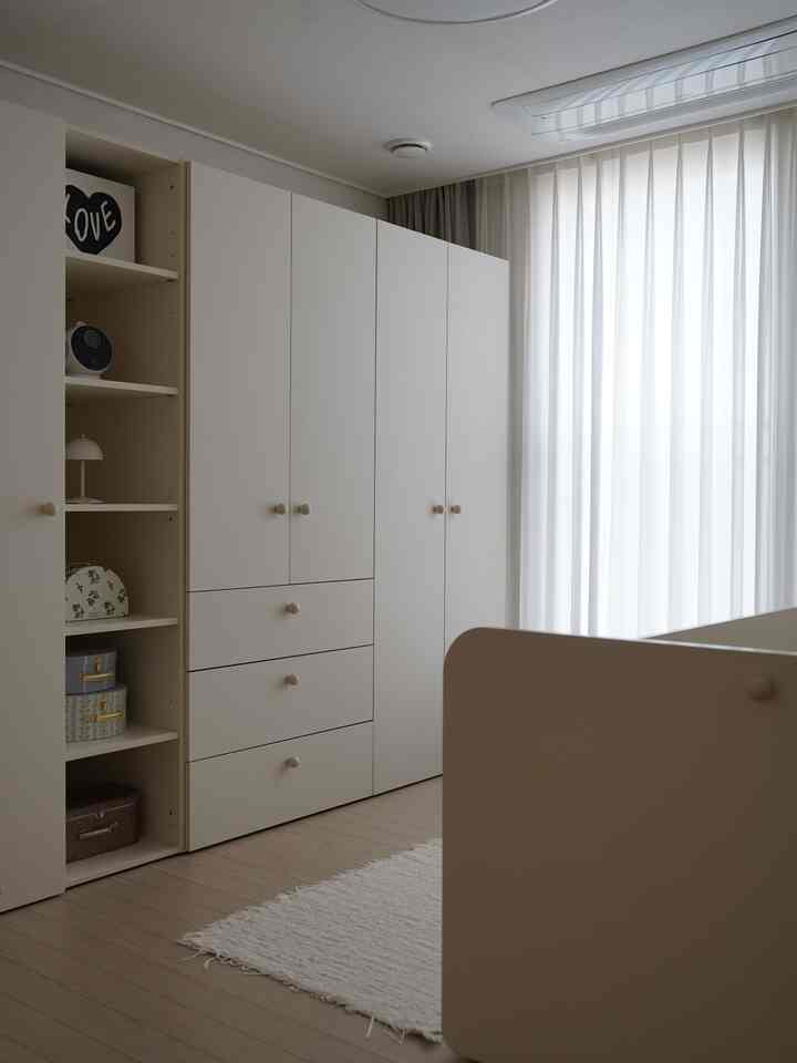 白とベージュのトーンの子供部屋、大きなワードローブとカーテン、デイベッドの一部が配置されたシンプルで清潔感のある空間
