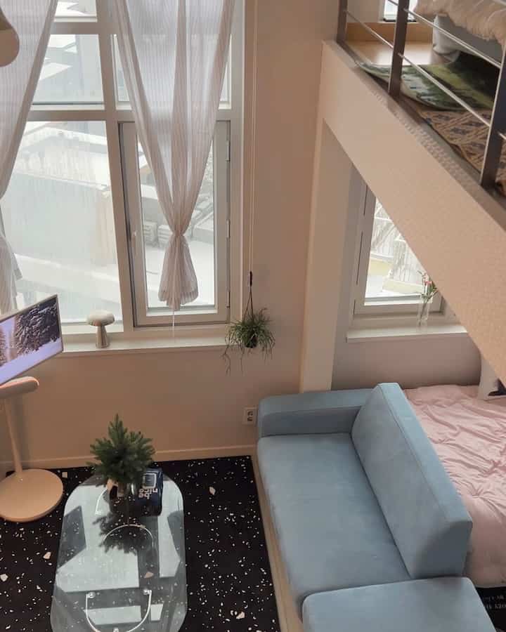 白と黒のモノトーンが基調の複層ワンルームリビング、淡い青のソファと透明なコーヒーテーブル、落ち着いたラグと植物が配置された雰囲気あるインテリア