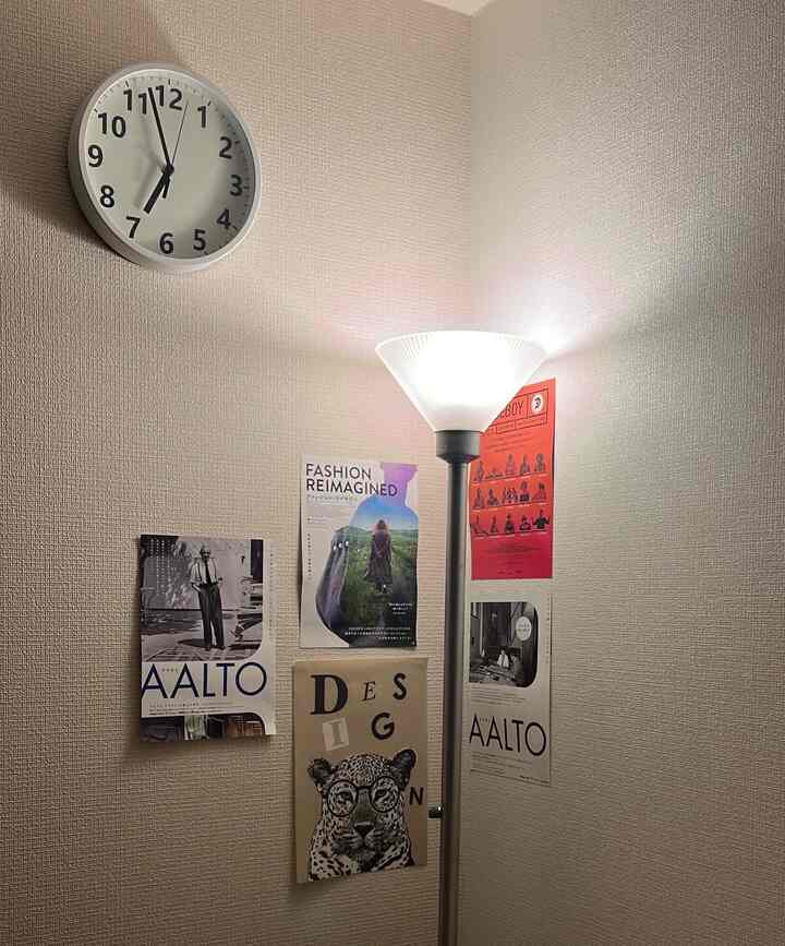 White-toned corner space featuring a wall clock, floor lamp, and various posters arranged in a simple setting